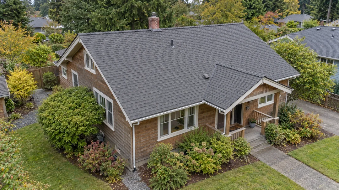 Low oblique aerial view of a cedar-sided Pacific Northwest bungalow, showing its asphalt-shingle gable roof, chimney and surrounding garden.